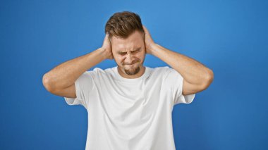 Sakallı, beyaz gömlekli, sıkıntılı genç bir adam, kulaklarını mavi bir arka plana kapatıyor. Hayal kırıklığını ya da gürültü izolasyonunu gösteriyor..