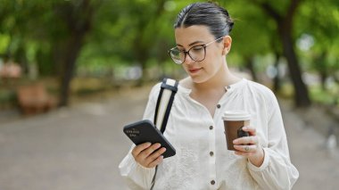 Genç ve güzel İspanyol kadın akıllı telefon kullanıyor. Parkta kahve içiyor.