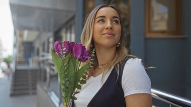 Genç, güzel, sarışın bir kadın bir şehir caddesinde dikilirken elinde mor lale buketi tutuyor. Açık havada çekici ve neşeli bir görünüm sergiliyor..