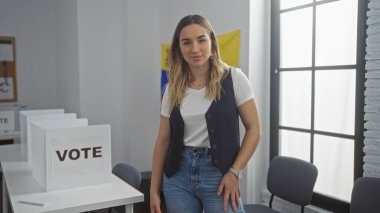 Güzel sarışın bir kadın, Venezuela 'da bir seçim koleji odasında duruyor, arka planda bayraklar ve pankartlarla kapalı bir yerde oy kullanma alanı gösteriyor..
