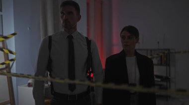 Ciddi bir adam ve kadın kapalı bir suç mahallini araştırıyor, uyarı bandıyla işaretlenmiş, şüphe ve soruşturmayı yansıtıyor..