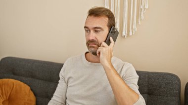 Modern bir oturma odasında sakallı, orta yaşlı bir adam düşünceli ve nişanlı görünüyor..