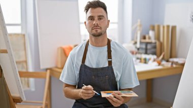 Önlük takan sakallı yakışıklı genç bir adam bir sanat stüdyosunda palet tutuyor.