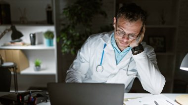 Laboratuar önlüklü, düşünceli bir erkek doktor kliniğinde geç saatlere kadar çalışıyor, dizüstü bilgisayar kullanıyor ve gözlük takıyor..