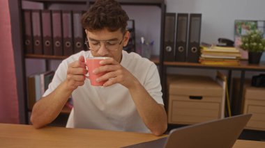 Ofiste kahve içen Latin kökenli genç adam, dizüstü bilgisayarı ve kitaplıklarla çevrili bir masada otururken bir anlık yansımayla yakalandı..