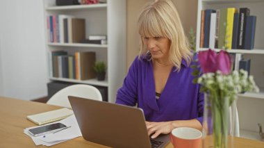 Özenle hazırlanmış sarışın kadın, arka planda bitki dekoru olan bir ofiste dizüstü bilgisayarla çalışıyor..