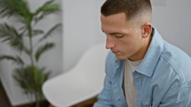 Açık mavi gömlekli yakışıklı genç adam arka planda bulanık bir bitkiyle modern bir odada düşünceli bir şekilde oturuyor.