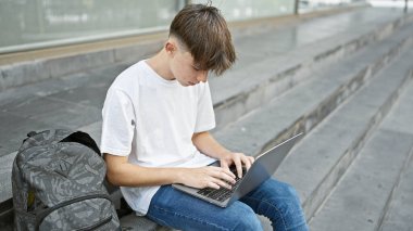 Genç beyaz erkek öğrenci şehir kampüsünün merdivenlerinde dizüstü bilgisayar kullanıyor..