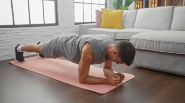 Formda bir adam modern oturma odasında pembe bir paspas üzerinde tahta egzersizi yapar. İçerde sağlıklı bir yaşam biçimi sergiler..