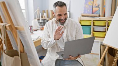 Gri sakallı, gülümseyen olgun bir adam sanat stüdyosundaki bir bilgisayardan video görüşmesi yapıyor..