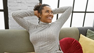 Rahat, kıvırcık saçlı, rahat bir oturma odasında gülümseyen Afro-Amerikalı kadın..