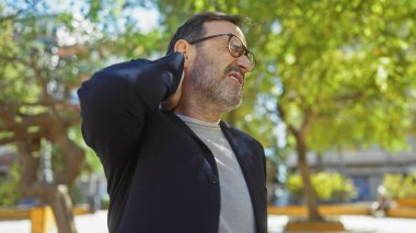 Sakallı, olgun bir adam güneşli bir parkta boyun ağrısı çekiyor.
