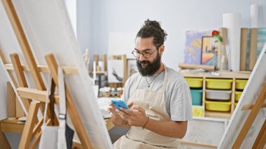Önlüklü sakallı genç bir adam resim sehpası ve tablolarla çevrili bir sanat stüdyosunda akıllı telefon kullanıyor..