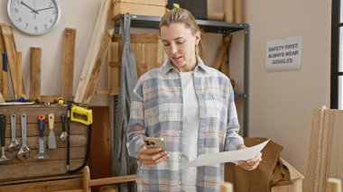 Odaklanmış bir kadın, marangozluk atölyesinde gazete okuyor ve elinde akıllı bir telefon tutuyor. Etrafı alet ve kerestelerle çevrili..