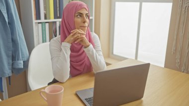 Tesettürlü düşünceli bir kadın ofis masasında oturur ve pencerenin yanındaki laptopundan gözünü ayırmaz..