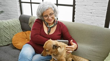 Gri saçlı, gülümseyen olgun bir kadın evde bir kanepede otururken köpeğini okşar..