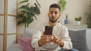 Yakışıklı bir İspanyol adam oturma odasında akıllı telefon kullanarak oturur. Etrafı iç mekan bitkileri ve rahat dekor ile çevrilidir. Modern ev hayatının bir anını yakalar..