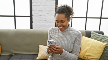 Şık bir oturma odasında rahat bir kanepede akıllı telefon kullanan kıvırcık saçlı, gülümseyen genç bir kadın..