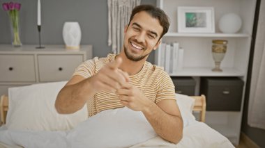 Sakallı, gülümseyen bir İspanyol genç adam iyi ışıklandırılmış bir yatak odasının içinde kameraya bakıyor ve ilgi çekici ve davetkar bir sahne yaratıyor..