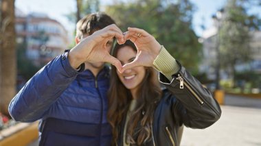 Sevgi dolu bir çift güneşli bir parkta elleri ile bir kalp şekli oluşturur. Açık havada sevgi, romantizm ve neşe gösterirler..