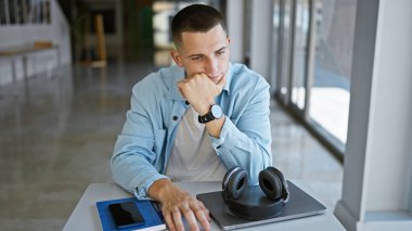 Üniversite kütüphanesinde laptopu, kulaklığı ve akıllı telefonuyla okuyan düşünceli bir genç..