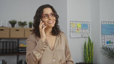Ofisteki genç, çekici İspanyol kadın, telefonda gülümsüyor ve konuşuyor, arka planda işyeri dekorasyonu ve bitkileri var..