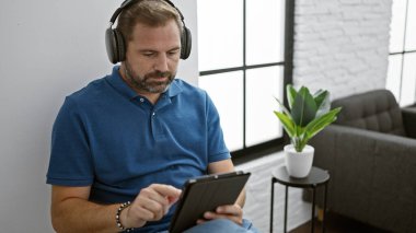 Gri saçlı, kulaklık takan yakışıklı, olgun bir adam modern ev ortamında tablet kullanıyor..