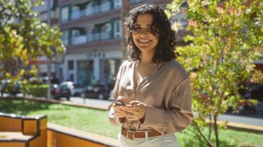 Güzel esmer, İspanyol bir kadın şehir parkında gülümsüyor, güneşli bir günde elinde akıllı bir telefon tutuyor..