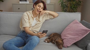Genç, beyaz bir kadın evcil köpeğiyle rahat bir oturma odasında dinleniyor telefonuna bakarken gülümsüyor..