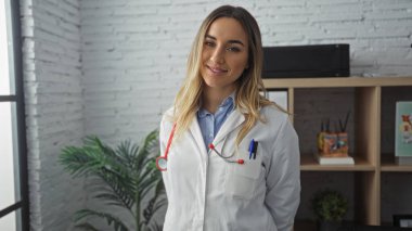Modern bir klinik çalışma odasında steteskopu olan beyaz önlüklü güzel sarışın bayan doktor..