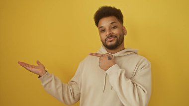 Yakışıklı siyah bir adam sarı arka planda avucunun içini göstererek dostça bir ifade sergilemeyi ya da sunmayı öneriyor..