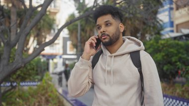 Şehir parkının dışında yeşil yeşillik ve şehir geçmişiyle telefonda konuşan günlük kıyafetli Afrikalı Amerikalı bir adam..