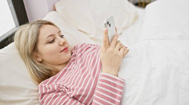 Sarışın kadın rahat bir yatak odasında akıllı telefonuyla dinleniyor.