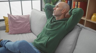 Evde gri bir kanepeye uzanmış sakallı, rahat kel bir adam. Düşünceli ve rahat görünüyor..