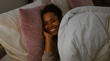 A young woman with curly hair smiles warmly while lying in bed, exuding coziness in an indoor bedroom setting. clipart