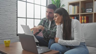 İspanyol bir adam ve kadın, yakınlarda bir dizüstü bilgisayar ve fincanla birlikte evdeki bir kanepede belgeleri inceliyor..