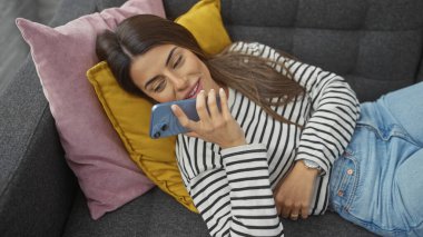 Genç bir İspanyol kadın renkli yastıklarla kanepede yatarken gülümsüyor, rahat bir evde akıllı telefonunu kullanıyor..