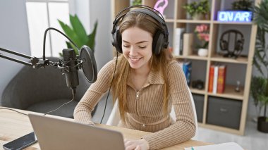 Genç sarışın bir kadın radyo stüdyosunda dizüstü bilgisayarla çalışmaktan, kulaklık takmaktan ve mikrofon başında gülümsemekten hoşlanıyor..