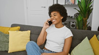 Gülümseyen Afrikalı Amerikalı kadın evde bir kanepede dinlenirken telefon konuşmasından zevk alıyor.