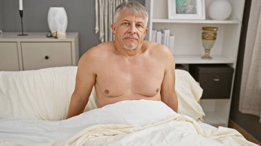 Gri saçlı orta yaşlı bir adam, zevkli dekore edilmiş bir yatak odasında, bir yatakta üstsüz oturur ve evdeki rahat ya da içe dönük bir anı canlandırır..