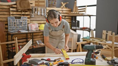 Odaklanmış genç bir kadın, iyi donanımlı bir marangozluk atölyesinde odun ölçer.