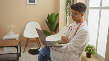 Laboratuvar önlüğü ve steteskop giyerek klinikte dizüstü bilgisayar kullanan ve sandalyeye oturan İspanyol bir adam..