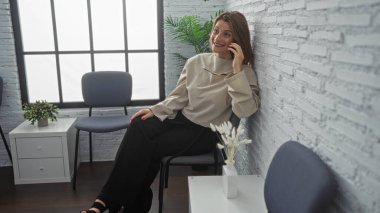 Genç, çekici, sarışın, beyaz bir kadın bekleme odasının koridorunda oturuyor, gülümsüyor ve telefonuyla konuşuyor, arka planda modern iç dekorasyon ve bitkiler var..