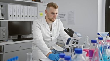 Genç beyaz bir adam, bilimsel ekipmanlarla çevrili modern bir laboratuvar ortamında mikroskoba odaklanıyor..