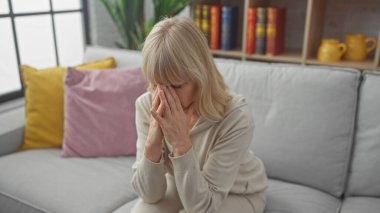 Sıkıntılı sarışın bir kadın evdeki koltukta oturur, elleri yüzünü kapatır, üzüntü ve stres hisseder..