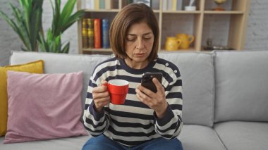 Orta yaşlı, çizgili kazaklı bir İspanyol kadın kapalı bir koltukta oturmuş akıllı telefonunu kontrol ederken kahvesini yudumluyor..