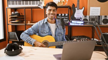 Yakışıklı, genç bir İspanyol, mikrofon ve dizüstü bilgisayarı olan bir müzik stüdyosunda gitar çalıyor.