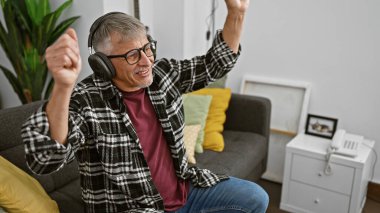 Kulaklıklı neşeli bir son sınıf öğrencisi modern bir oturma odasındaki koltukta otururken kutlama yapıyor.