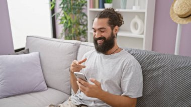 Oturma odasındaki kanepede akıllı telefon kullanarak gülümseyen sakallı adam evdeki rahat ve modern yaşam tarzını resmediyor..