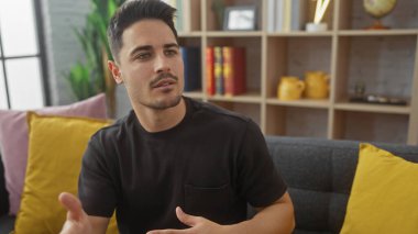 Siyah gömlekli, sakallı, modern bir oturma odasında konuşan ve el kol hareketi yapan yakışıklı bir İspanyol.